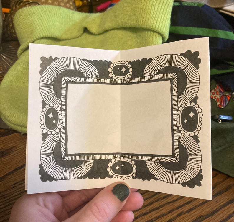 Puede incluir: Una tarjeta blanca plegada con un borde decorativo en blanco y negro. El borde presenta un patr&oacute;n repetitivo de l&iacute;neas curvas y formas ovaladas. La tarjeta se sostiene en una mano con esmalte de u&ntilde;as verde oscuro.