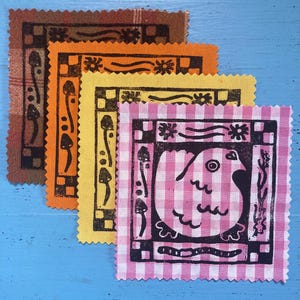 May include: Four square fabric patches with pink-and-white gingham, yellow, orange, and brown plaid patterns. Each patch features a black ink block print of a bird or mushrooms within a decorative border of squares, flowers, and wavy lines.
