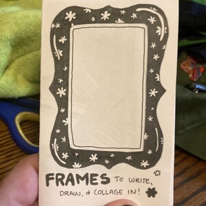 Puede incluir: Un marco de papel rectangular con un borde negro decorado con peque&ntilde;as flores blancas en forma de estrella. El texto "FRAMES TO WRITE, DRAW, & COLLAGE IN!" est&aacute; impreso en la parte inferior del marco. El marco est&aacute; sostenido por una persona.