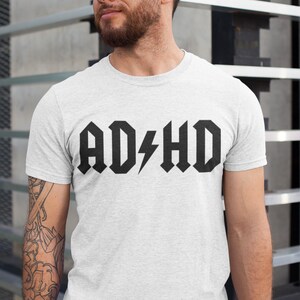 Pode incluir: Uma camiseta branca com um gr&aacute;fico preto que diz "AD/HD" em uma fonte de rock and roll.