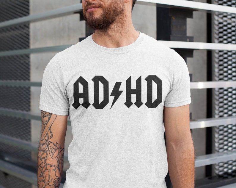 Pode incluir: Uma camiseta branca com um gr&aacute;fico preto que diz "AD/HD" em uma fonte de rock and roll.