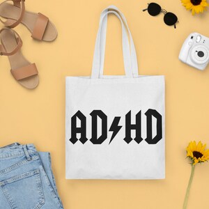 Pode incluir: Sacola de tecido branca com texto preto que diz "AD/HD" em uma fonte de rock and roll com um raio no lugar da barra obl&iacute;qua. A sacola est&aacute; sobre uma superf&iacute;cie amarela com sand&aacute;lias, &oacute;culos de sol, uma c&acirc;mera e girass&oacute;is.