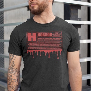 Puede incluir: Camiseta gris oscuro con un gr&aacute;fico rojo que dice "HORROR" con una lista de elementos de terror y goteo de sangre. La camiseta tiene cuello redondo y mangas cortas.