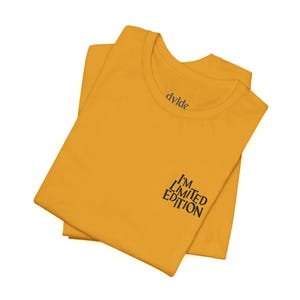 Peut inclure: T-shirt jaune moutarde plié, avec l'inscription noire "I'M LIMITED EDITION" en bas à droite. Le nom de la marque "dvlde" est imprimé au dos du col. Le tissu semble doux.