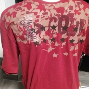 May include: A red short-sleeve t-shirt featuring a tan camouflage-style pattern across the upper back and shoulders. The design includes the text "X-COL" with the "X" embellished in silver rhinestones and dark brown star graphics below.