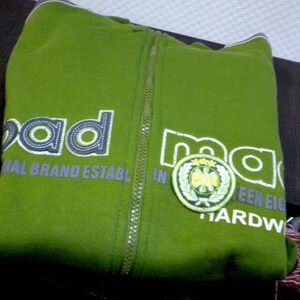May include: A green zippered hoodie with the word "mad" printed on the front in white and gray lettering. The hoodie also has a green and white patch with a logo on the right side of the front. The text "National Brand Establ. In ...teen Eigh... Hardw..." is printed on the front of the hoodie.