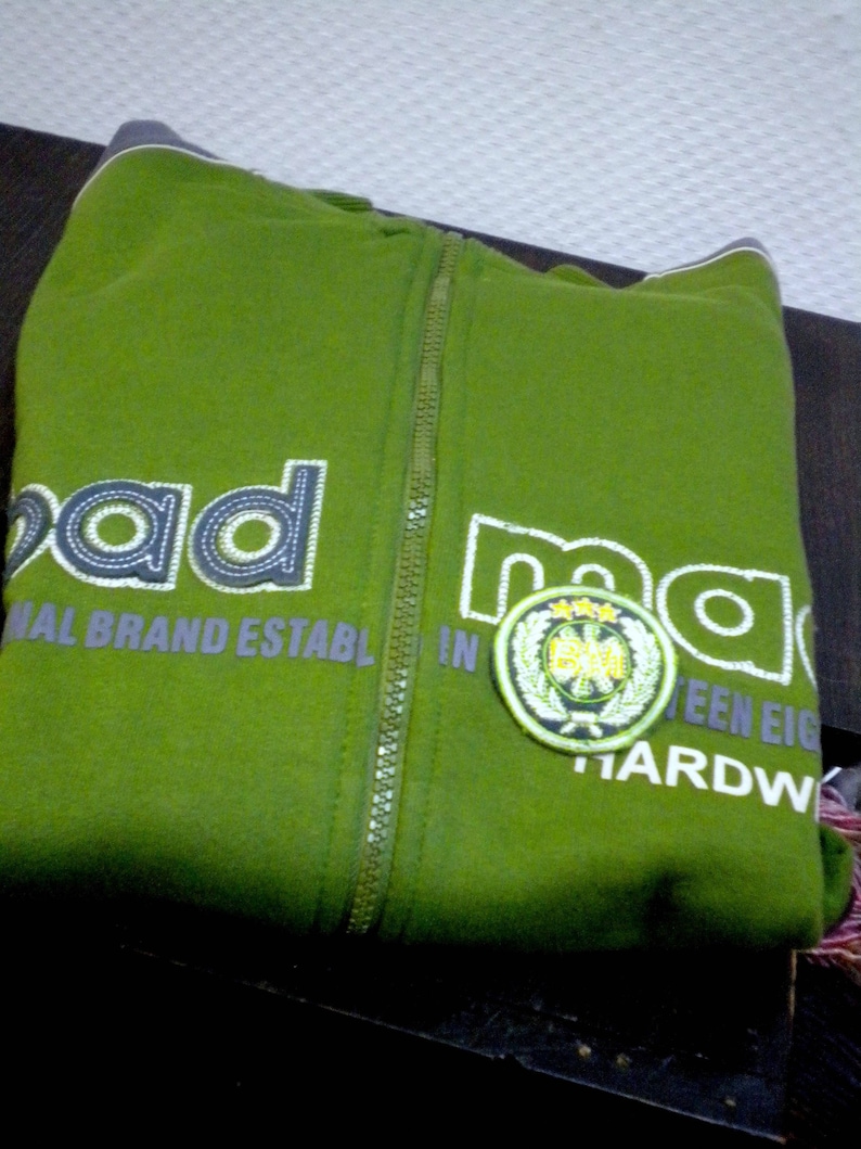 May include: A green zippered hoodie with the word "mad" printed on the front in white and gray lettering. The hoodie also has a green and white patch with a logo on the right side of the front. The text "National Brand Establ. In ...teen Eigh... Hardw..." is printed on the front of the hoodie.