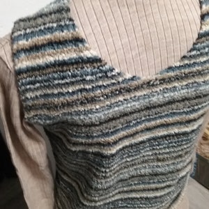 May include: A textured, sleeveless sweater in shades of blue, grey, and brown. The sweater is layered over a beige turtleneck. The design features horizontal stripes, creating a cosy and stylish look. The garment appears to be handmade.