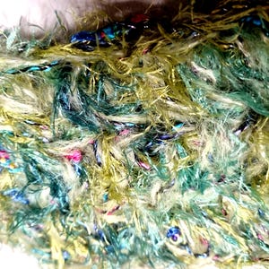 Könnte beinhalten: Nahaufnahme von strukturiertem Garn in Grün-, Gelb- und Weißtönen. Das Garn hat ein flauschiges, fransiges Aussehen mit eingewebten blauen, rosa und schwarzen Strähnen. Das Garn wirkt weich und könnte zum Stricken oder für andere Bastelarbeiten verwendet werden.
