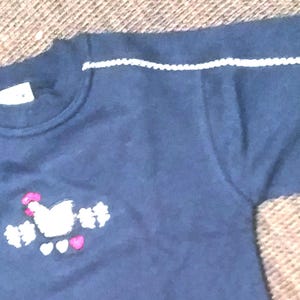 Op de afbeelding: Marineblauw shirt met lange mouwen en witte biezen langs de schouders en mouwen. Het shirt heeft een geborduurd ontwerp van een witte kip met roze accenten en hartjes. Een label is zichtbaar op de kraag.