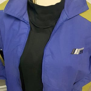 May include: A blue and yellow vintage jacket with a black turtleneck underneath. The jacket has a zipper closure and a logo on the chest. The jacket is displayed on a mannequin.