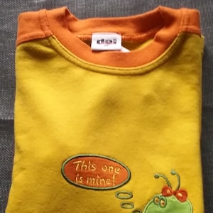 May include: Yellow t-shirt with an orange trim around the neck. The shirt has an embroidered green bug with red accents and a speech bubble that says "This one is mine!"