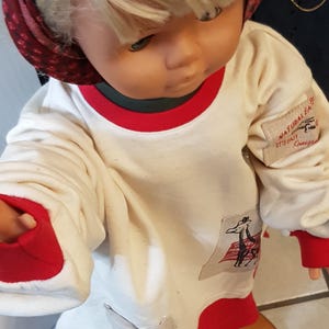 May include: A vintage doll with blonde hair and blue eyes, wearing a white long-sleeved shirt with red trim. The shirt features patches with illustrations and text. The doll is wearing a red and patterned headscarf.
