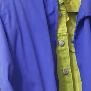 May include: A blue and green jacket with a zipper and button closure. The jacket has a blue exterior with green and yellow accents. The buttons are silver. The jacket is hanging on a hanger.
