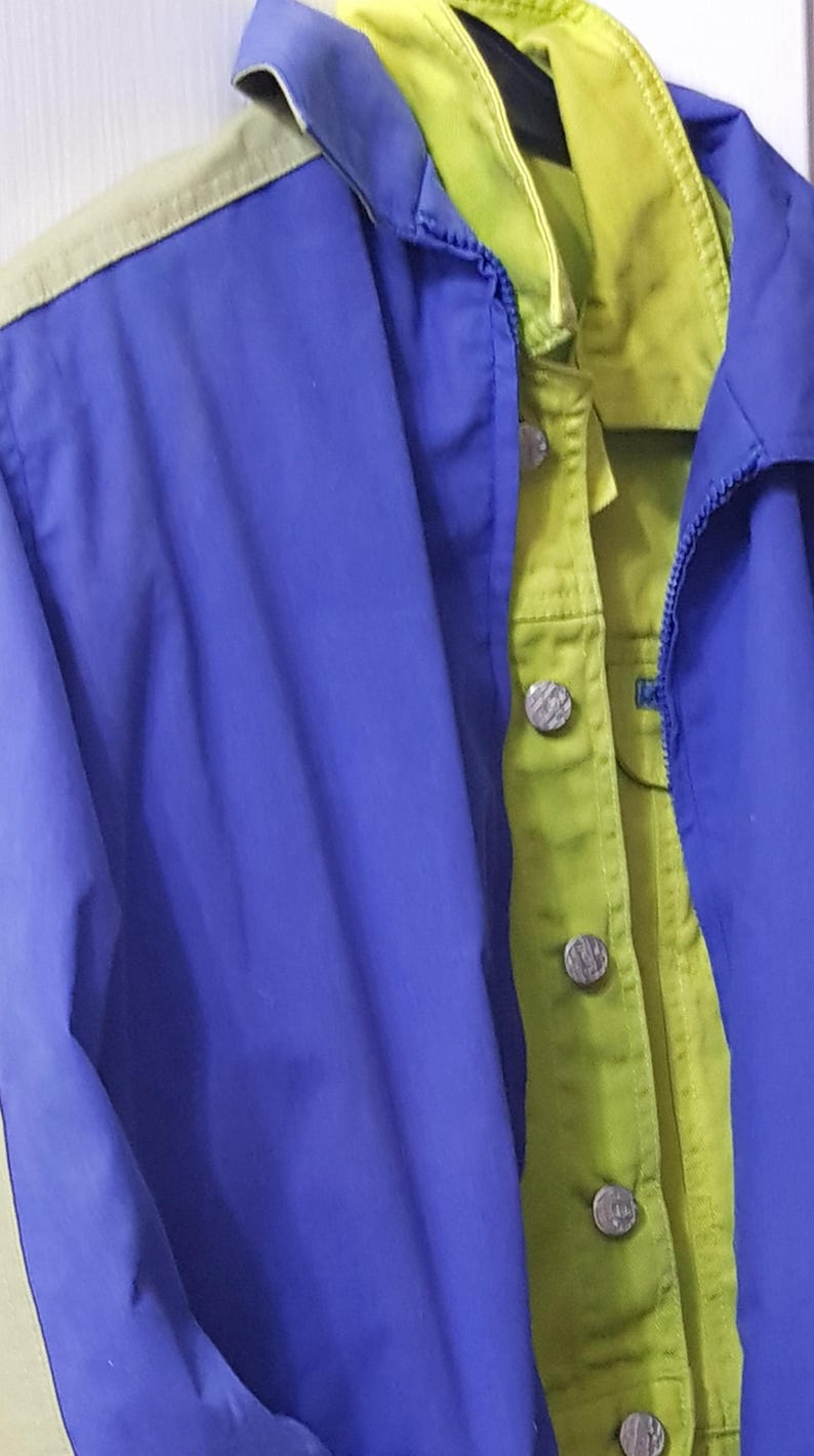 May include: A blue and green jacket with a zipper and button closure. The jacket has a blue exterior with green and yellow accents. The buttons are silver. The jacket is hanging on a hanger.