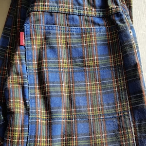 May include: Blue plaid pants with a red tag on the back pocket. The plaid pattern includes blue, green, yellow, and orange lines. The pants are folded.