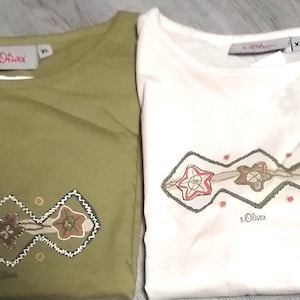 Könnte beinhalten: Zwei T-Shirts, eines olivgrün und eines weiß, werden gezeigt. Jedes Shirt hat einen Rundhalsausschnitt und ein gesticktes Design mit Stern- und Blumenmotiven in Rot-, Braun- und Grüntönen. Die Marke "s.Oliver" ist sichtbar.