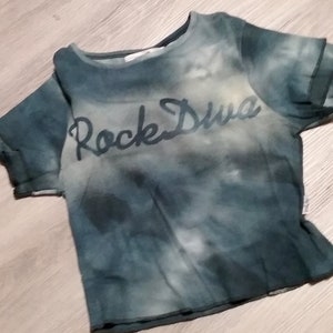 May include: A short-sleeved, tie-dye style t-shirt in shades of blue and gray. The words "Rock Diva" are printed across the front in a dark, cursive font. The shirt has a crew neck and a slightly distressed look.