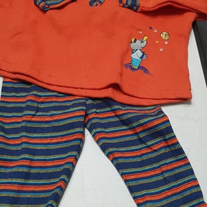 Puede incluir: Un conjunto de bebé de dos piezas en naranja y azul. La parte superior es un jersey de manga larga con cremallera y un pequeño diseño de animal bordado. Los pantalones son de rayas azules, naranjas y amarillas.