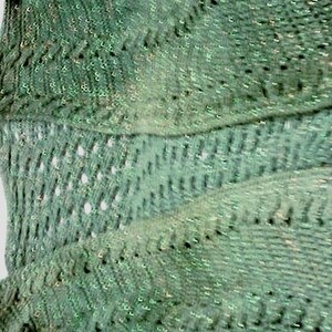 May include: A close-up of a green knitted shawl with a textured, openwork design. The shawl has a shimmering, iridescent quality, with a pattern of small holes and raised stitches. The overall effect is soft and flowing.