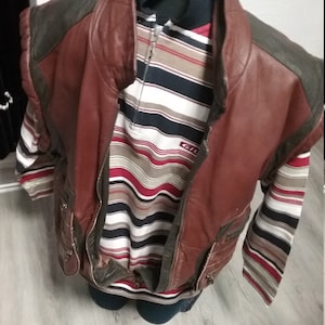 Puede incluir: Un chaleco de cuero marrón con detalles en los hombros de color verde oscuro se superpone a una camisa a rayas con rayas rojas, blancas y negras. El chaleco tiene cierre de cremallera y bolsillos. La camisa tiene mangas largas.