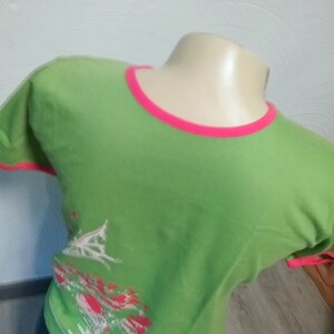 Könnte beinhalten: Ein lindgrünes Kurzarm-T-Shirt mit einem leuchtend pinkfarbenen Rundhalsausschnitt und passenden Ärmelabschlüssen. Auf der unteren linken Seite befindet sich ein Grafikdruck in Weiß- und Rottönen, der ein stilisiertes baumartiges Motiv zeigt.