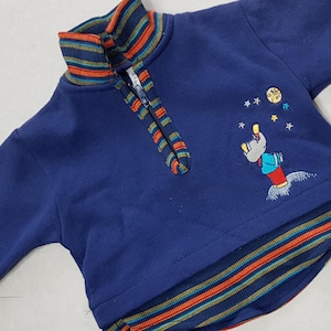 Op de afbeelding: Een blauw longsleeve sweatshirt met een gestreepte kraag en een rits. Het sweatshirt heeft een geborduurd ontwerp van een cartoonfiguur die naar een ster reikt.