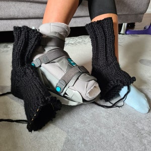 Könnte beinhalten: Eine Person trägt einen schwarzen gestrickten Beinstulpen an ihrem linken Bein und einen grauen Knöchelstiefel mit einem schwarzen Riemen. Die Person sitzt auf einem Sofa mit ausgestrecktem rechten Bein.