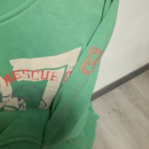 May include: Green long-sleeve sweatshirt with a white graphic print that says "Rescue" and a red patch on the sleeve that says "Mission Control".