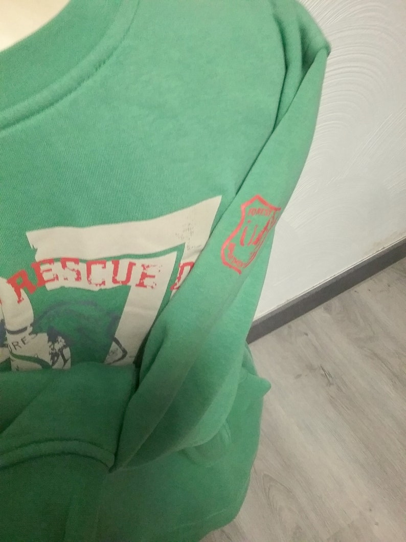 May include: Green long-sleeve sweatshirt with a white graphic print that says "Rescue" and a red patch on the sleeve that says "Mission Control".