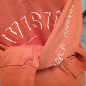 Op de afbeelding: Oranje longsleeve sweatshirt met witte tekst die "VISTA" op de voorkant en "VISTA" herhaald op de linkermouw.