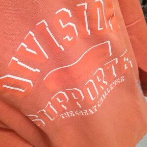 Op de afbeelding: Oranje sweatshirt met witte tekst die "Division" en "Support The Great Challenge" luidt.