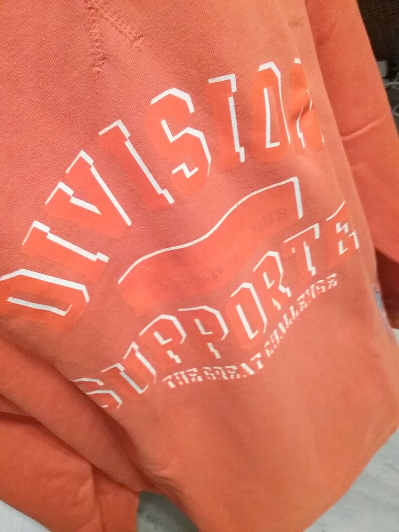 Op de afbeelding: Oranje sweatshirt met witte tekst die "Division" en "Support The Great Challenge" luidt.