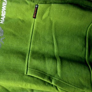 May include: Green hooded sweatshirt with a large graphic print on the front. The print includes the text "Original Brand Established in the Nineteen Eighty" and "Hardware". The sweatshirt has a kangaroo pocket on the front.