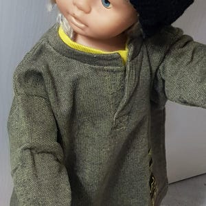 Könnte beinhalten: Eine Puppe trägt ein grünes Langarmshirt mit einem gelben Unterhemd und eine schwarze Strickmütze.