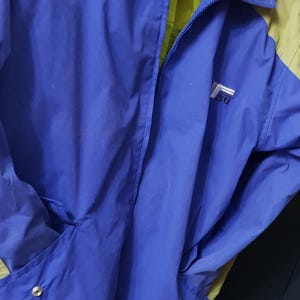 May include: A blue and green jacket with a zipper closure. The jacket has a logo on the chest and two pockets. The interior lining is green. The jacket is made of a lightweight material.
