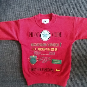 Könnte beinhalten: Ein rotes Sweatshirt mit langen Ärmeln, bestickt mit Text und Aufnähern. Der Text beinhaltet "PILOT CODE", "MESSAGE FROM C.6 DIVISION" und "TAKE YOUR POSITIONS". Die Aufnäher sind grün und goldfarben. Das Sweatshirt ist aus weichem Material.