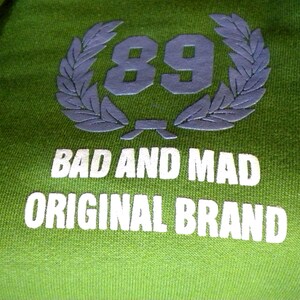 May include: Green sweatshirt with a white logo that says "Bad and Mad Original Brand" and a laurel wreath with the number 89 in the center.