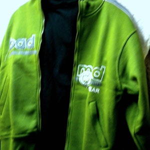 May include: A green zippered jacket with a black turtleneck underneath. The jacket has the word "rad" written on it in white and gray letters.
