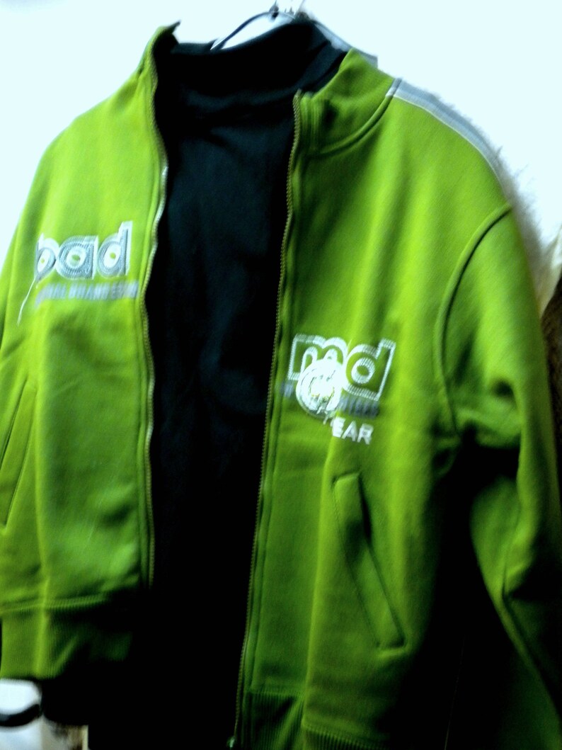 May include: A green zippered jacket with a black turtleneck underneath. The jacket has the word "rad" written on it in white and gray letters.