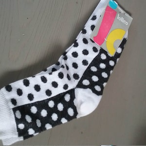 May include: A pair of crew-length socks featuring a black and white polka dot pattern. The socks are made of a knit fabric and include a branded tag at the top with the text jolico in white letters against a grey, pink, and yellow background.