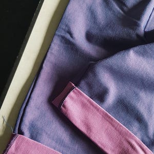 May include: A pair of purple sweatpants with a contrasting pink cuff. The pants are made of a soft fabric and have a drawstring waistband. The cuff is folded over, revealing the pink interior.