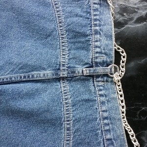 Può includere: Primo piano di un capo in denim blu con cuciture a vista e dettagli a catena color argento. Il capo presenta una serie di anelli e fibbie in metallo, con una catena che corre lungo il bordo. Il denim ha un aspetto sbiadito.