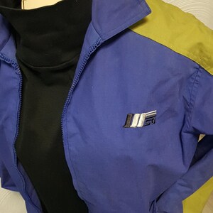 May include: A blue and yellow vintage jacket with a black turtleneck underneath. The jacket has a zipper closure and a small logo on the chest. The jacket is made of a lightweight material.