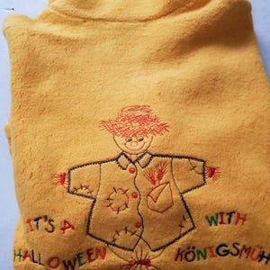 May include: A yellow fleece pullover with an embroidered scarecrow design. The text "It's a Halloween Party with Königsmühl" is embroidered on the front.