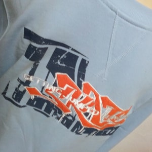 May include: Light blue sweatshirt with a graphic print of a red and white logo that says "Getting Stronger" in a stylized font.