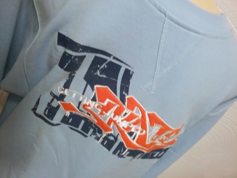 May include: Light blue sweatshirt with a graphic print of a red and white logo that says "Getting Stronger" in a stylized font.