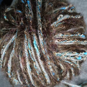 Op de afbeelding: Close-up van een bol garen met een mix van bruine, grijze en blauwe vezels. Het garen heeft een getextureerd uiterlijk met strengen metallic gouddraad. De bol is waarschijnlijk voor brei- of haakprojecten.
