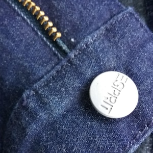 Könnte beinhalten: Nahaufnahme von dunkelblauem Denim-Stoff mit einem goldenen Reißverschluss und einem silbernen Knopf. Der Knopf ist rund und trägt den geprägten Schriftzug "ESPRIT". Das Bild zeigt die Details der Jeans.