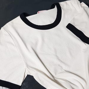 Può includere: T-shirt bianco a coste con finiture nere su colletto e maniche corte. La maglietta ha un design classico e sportivo. Il tessuto sembra essere un materiale morbido e strutturato.